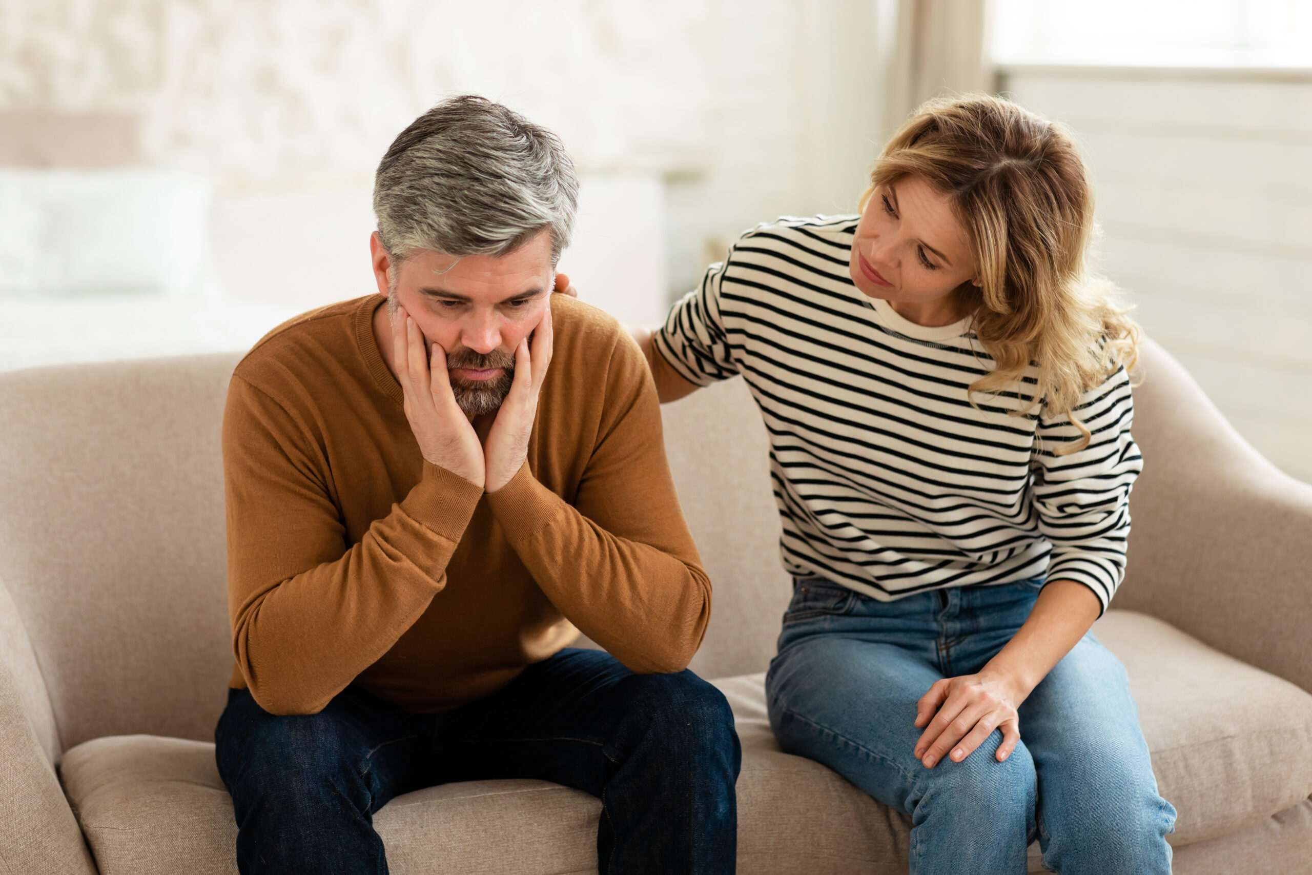 Middle Aged Wife Comforting And Supporting Depressed Husband Sitting On Couch At Home. Couple Struggling And Suffering Having Problems. Male Depression And Midlife Crisis Concept