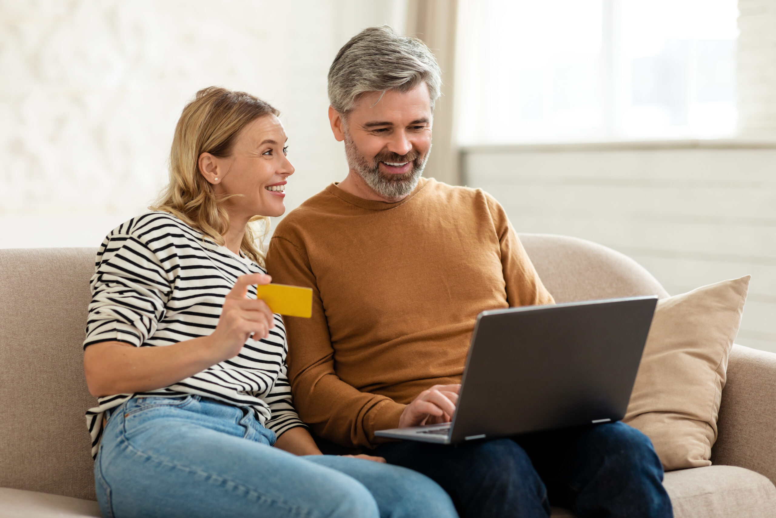 Online Shopping. Happy Middle Aged Couple Making Internet Payment Using Laptop Computer And Credit Card Sitting On Sofa At Home. Ecommerce, Technology And Consumerism